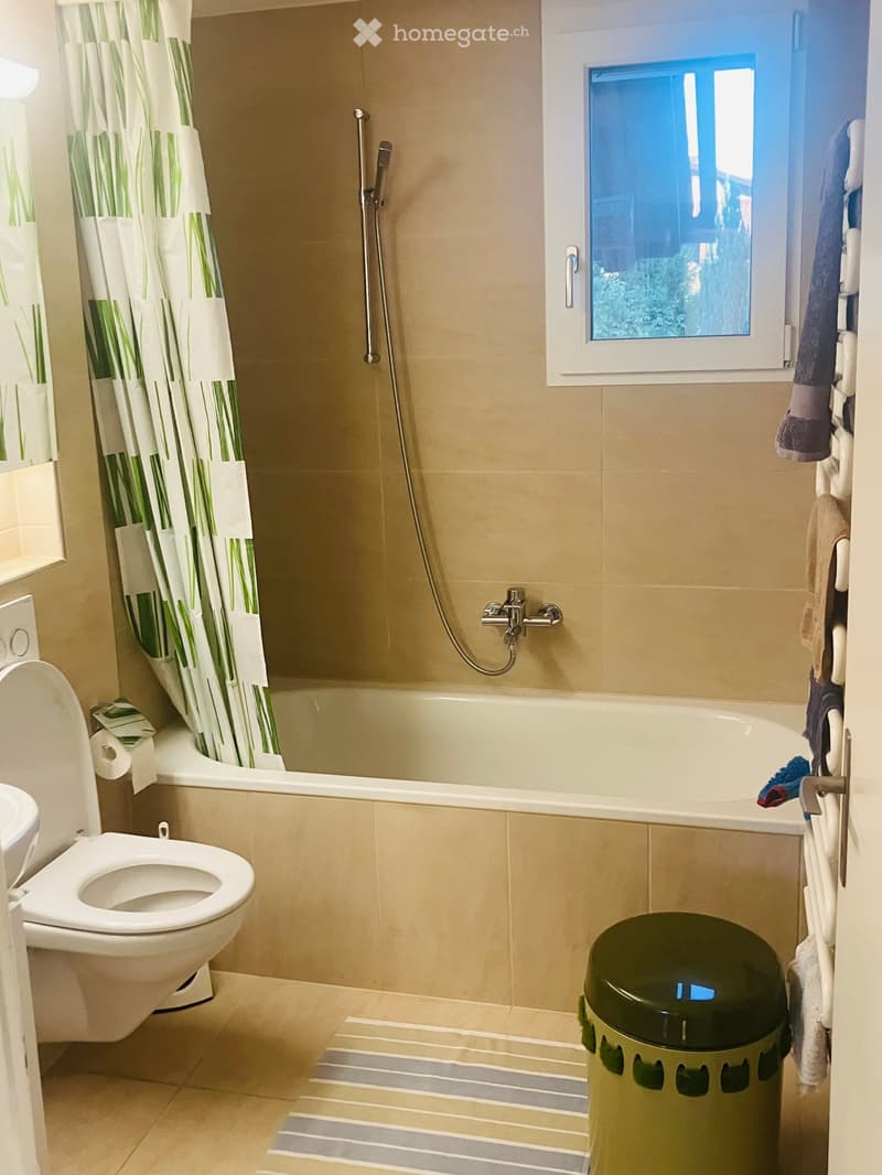 Modern bathroom with beige tiles, a bathtub with a shower curtain, a toilet, a towel rack, and a window offering a view of greenery.