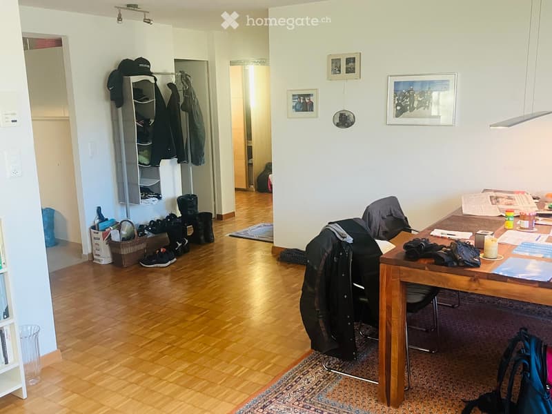 A furnished room with a wooden floor, a large wooden table with a chair, a coat rack with jackets and shoes, a bookshelf, and a small basket with items.