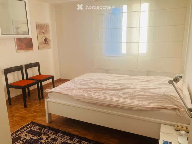 A small, well-lit bedroom with a white bed, two black chairs with orange seats, a nightstand with a lamp, and artwork on the walls.