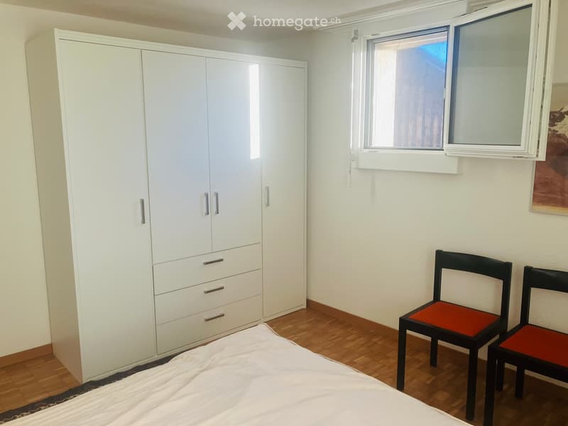 A modern bedroom with a white wardrobe, a bed with white bedding, and two black chairs with red cushions. The room features wooden flooring and a large window with white frames.