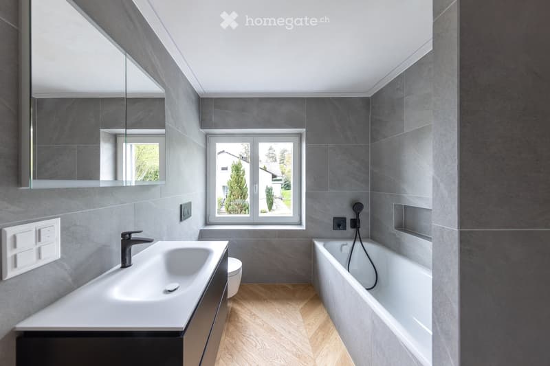 Modern bathroom with grey tiled walls, a white sink, a bathtub, a large mirror, and a window offering a view of the garden.