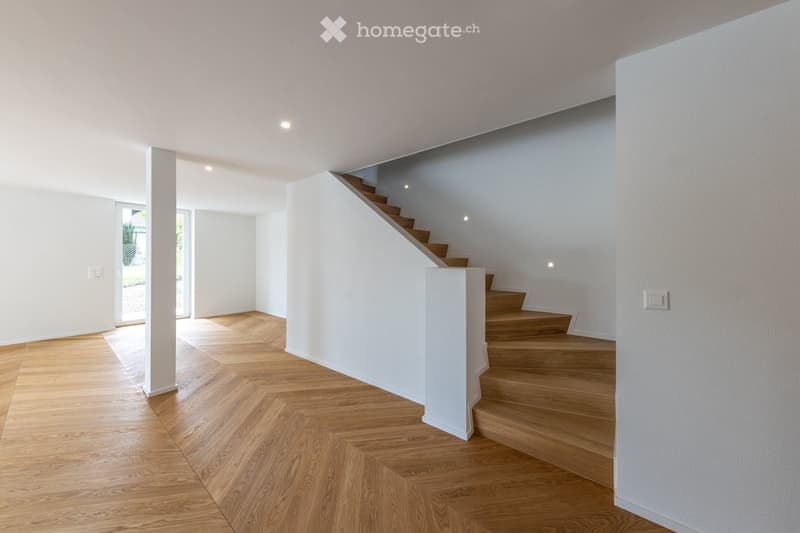 Bright, modern hallway with wooden flooring and white walls, featuring a staircase leading to an upper level and a door opening to an outdoor area.