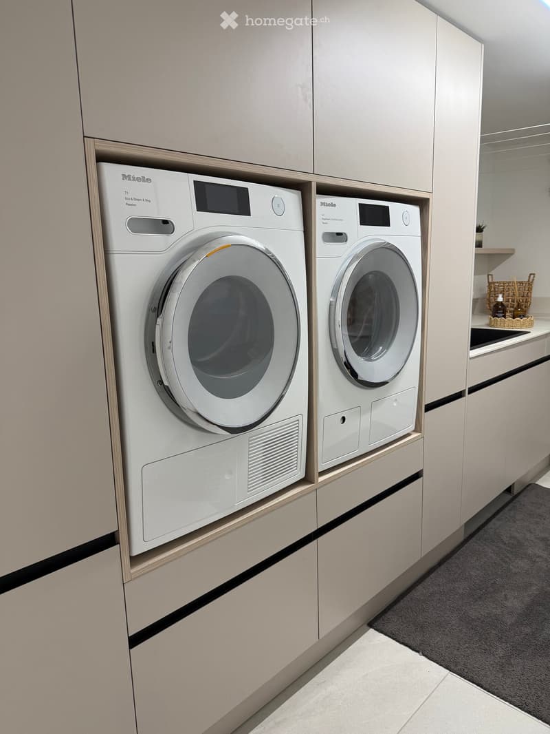 Modern bathroom with built-in Miele washing machine and dryer, light wood cabinetry, and a small shelf with decorative items.