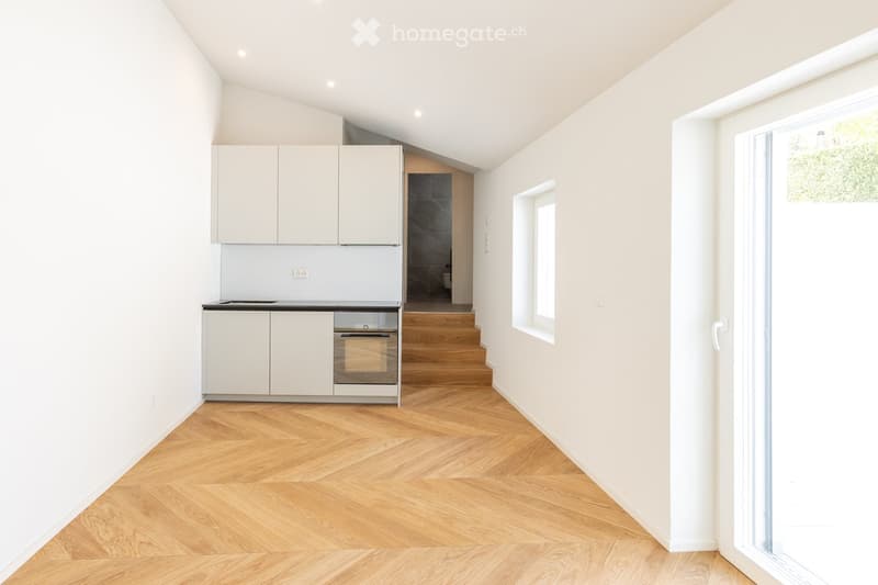 Modern, minimalist kitchen with white cabinets, a sink, and an oven. The room features light wooden flooring and white walls, with a small window and a view of a staircase leading to another level.