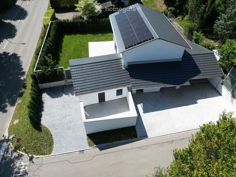 Modern two-story house with grey tiled roof, white walls, solar panels on one section, surrounded by a well-maintained garden and driveway.