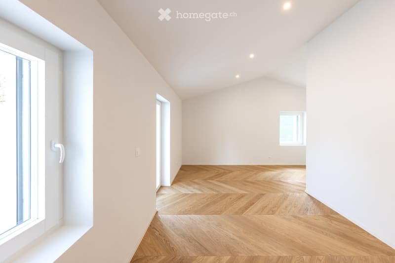Empty room with light-colored walls, wooden herringbone flooring, and large windows allowing natural light. No furniture or personal items are present.