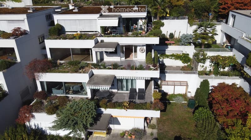 Aerial view of a modern white house with multiple levels, surrounded by lush greenery and trees. Features include large windows, terraces, outdoor seating, and a well-maintained garden.