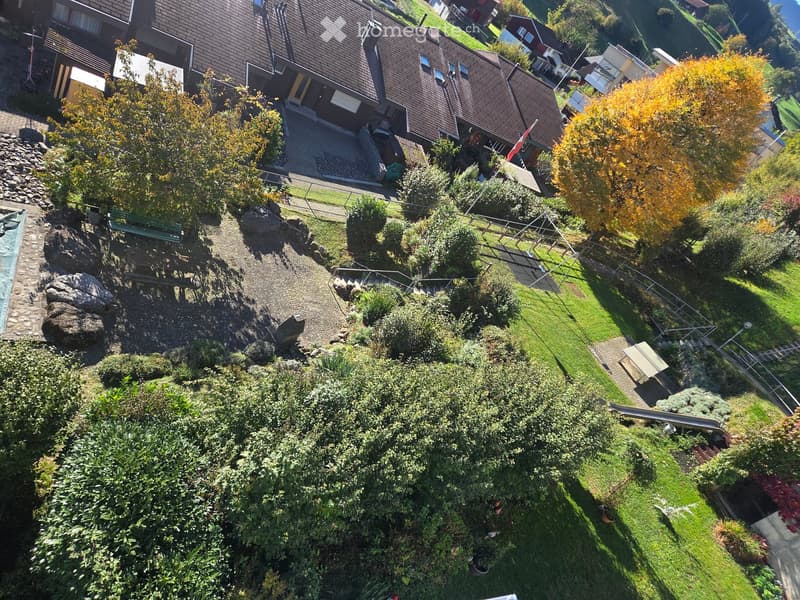 Aerial view of a landscaped garden with a swimming pool, green lawn, trees, and a small wooden deck. The garden is bordered by a stone pathway and includes a bench and a small staircase leading to a higher level.