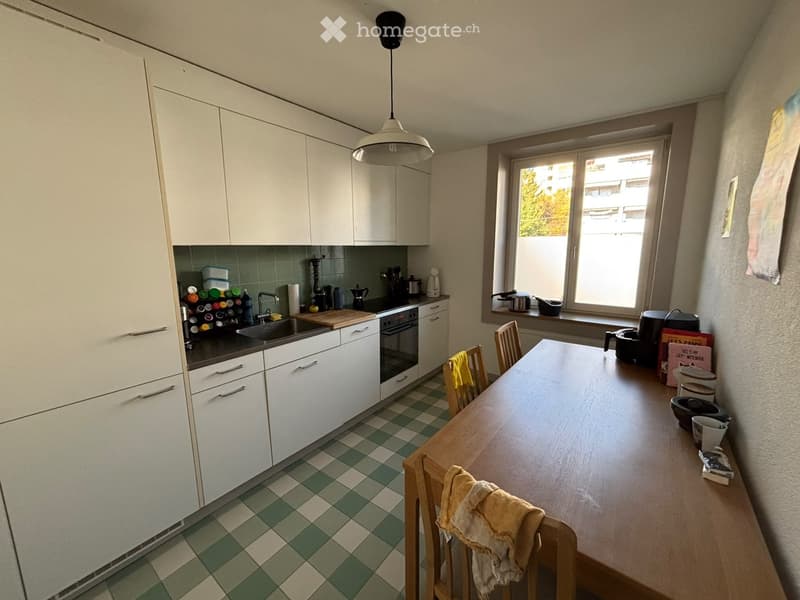 Modern kitchen with white cabinets, green tiled backsplash, and a wooden dining table with chairs. The kitchen features a sink, oven, and various appliances.