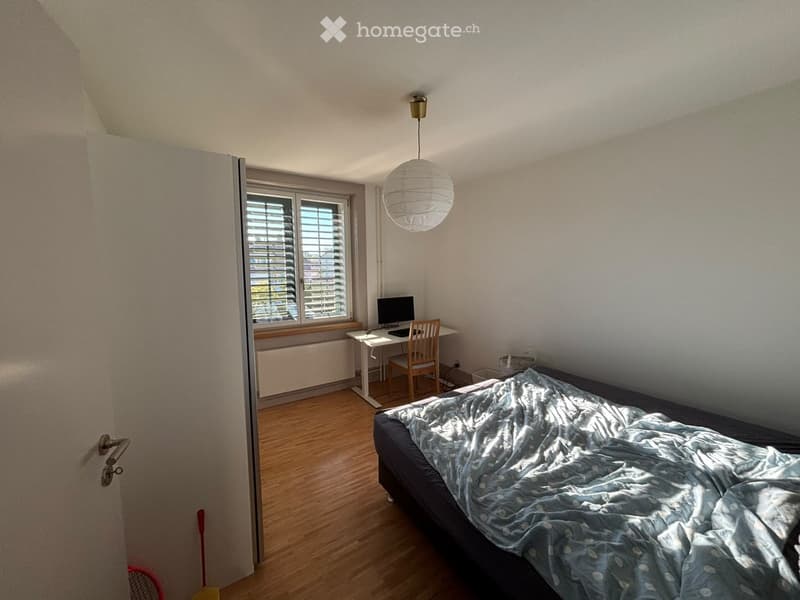 Cozy bedroom with wooden flooring, a bed with a blue patterned duvet, a desk with a computer and chair, and a hanging paper lantern light fixture.