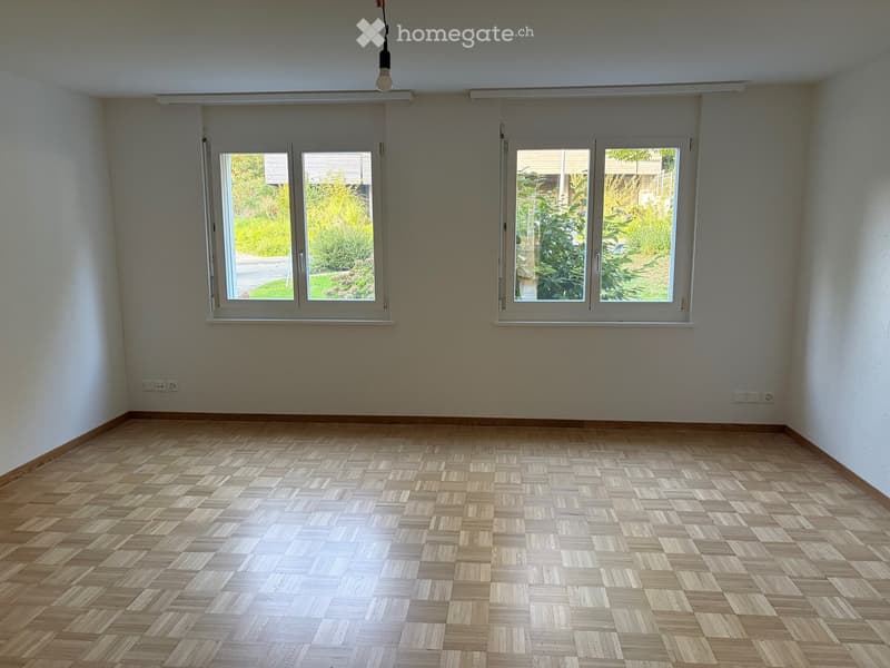 Empty room with wooden herringbone flooring, white walls, and large windows showing a garden view.