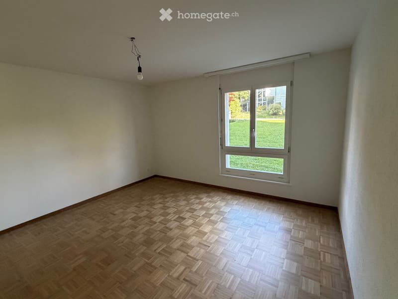 Empty room with wooden parquet flooring, white walls, and a large window with white frame showing a green outdoor view.