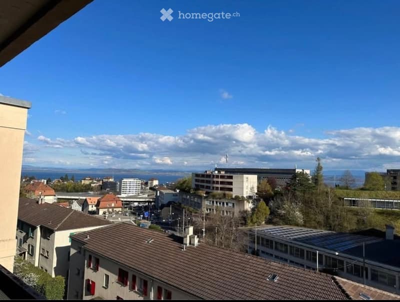 Panoramic cityscape view from a balcony, featuring rooftops, buildings, and distant water body under a clear blue sky.