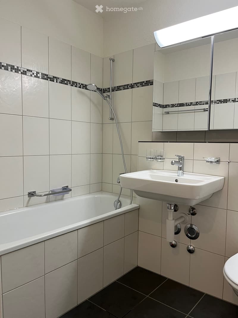 Modern bathroom with a bathtub, sink, and mirror. Features tiled walls and floor, with a chrome faucet and a glass shelf above the sink.