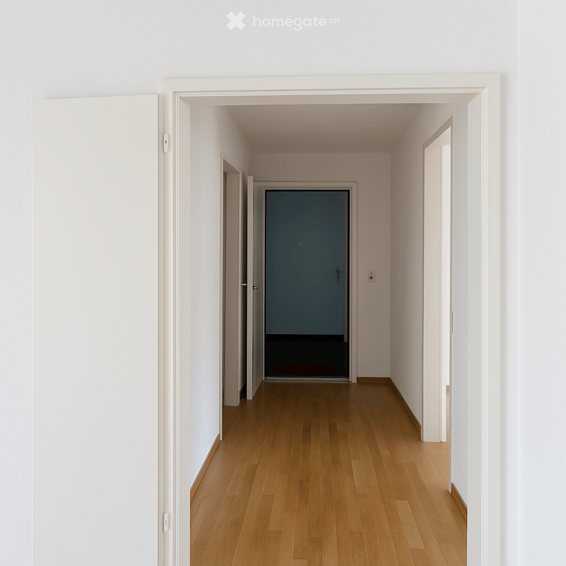 A clean, minimalist hallway with light wooden flooring and white walls. The corridor features several doors and a visible electrical outlet on the right wall.