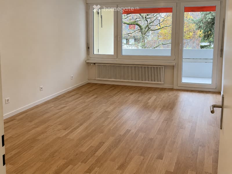 Empty room with light wooden flooring, white walls, and large windows with blinds. A radiator is visible under one window, and there are electrical outlets on the walls.