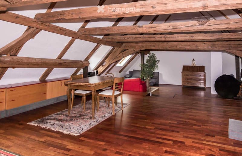 Spacious room with wooden beams, hardwood floor, dining table with chairs, sofa, ottoman, cabinet, and a plant.