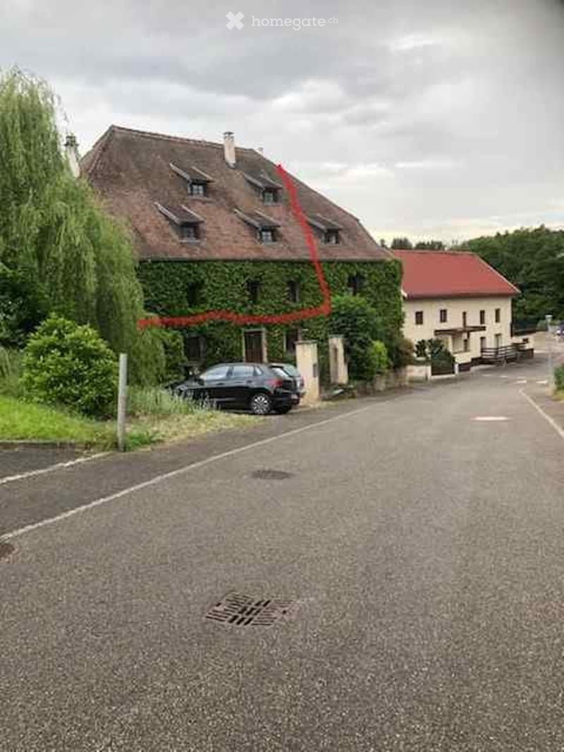 A two-story house with a tiled roof and ivy-covered walls, parked car in front, surrounded by greenery.