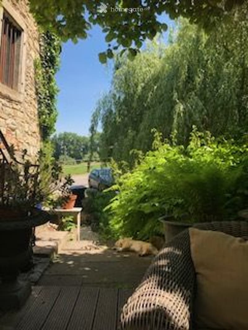 A serene garden area with a wooden deck, lush greenery, and a cozy seating area. The space features a wicker chair, a small table, and various potted plants, creating a tranquil outdoor retreat.