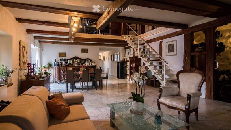 Spacious living and dining area with exposed wooden beams, a staircase leading to an upper level, a sofa, coffee table, armchair, dining table with chairs, a cabinet, and decorative items.