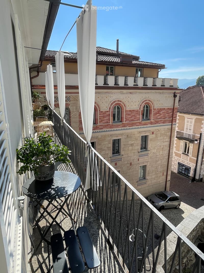 Balcony with a small round table, two chairs, and a potted plant, overlooking a historic building and a narrow street with a parked car.