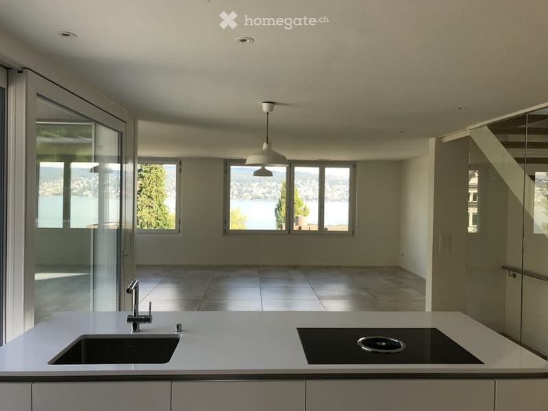 Modern kitchen with white countertops, a sink, faucet, and induction cooktop, large windows offering a scenic view of a lake and mountains.