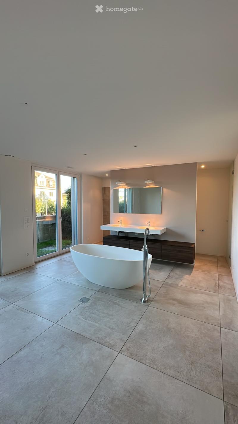 Modern bathroom with a freestanding bathtub, double sink vanity, large mirror, and sleek faucet. The space features large tiles and a sliding glass door leading to an outdoor area.
