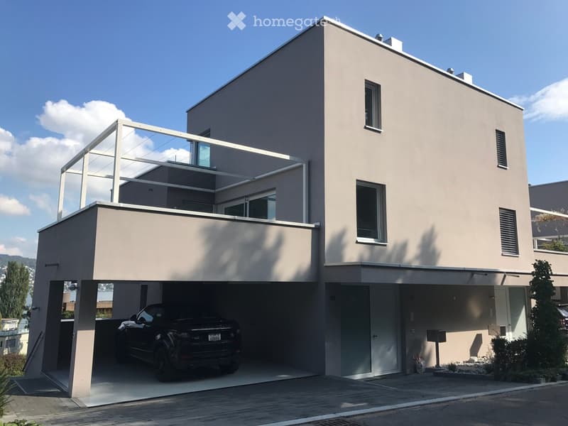 Modern two-story house with a light beige facade, large windows, and a spacious balcony on the upper level. The ground floor features a covered garage with a black SUV parked inside.