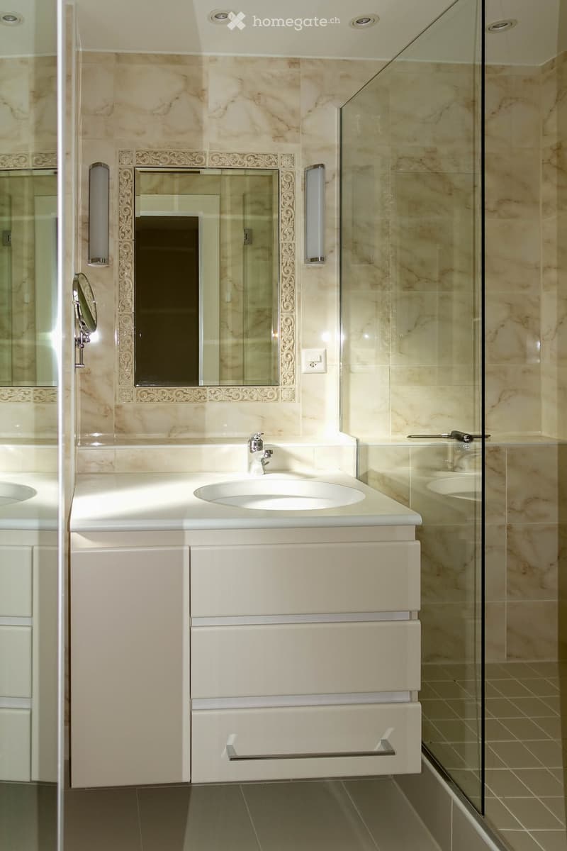 Modern bathroom with marble walls, a double sink vanity, a large mirror with ornate frame, and a glass-enclosed shower area.