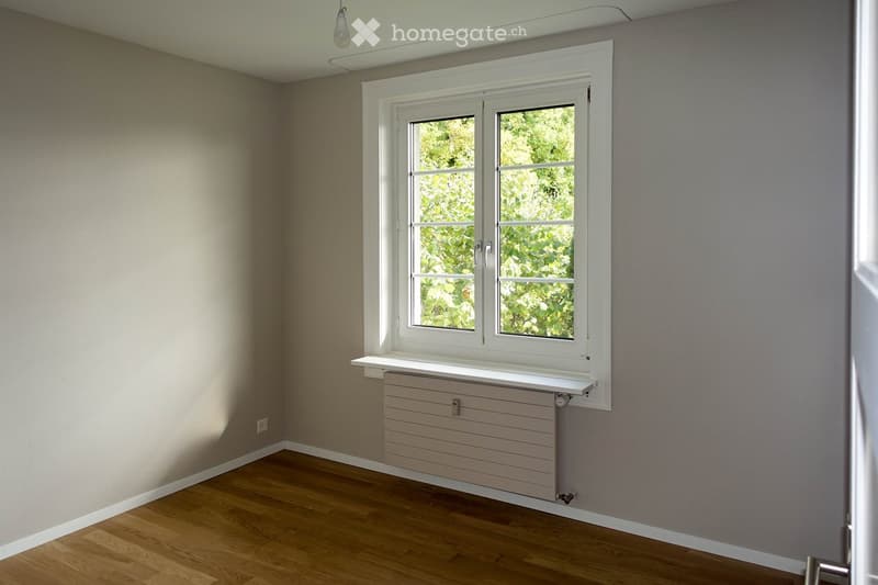 Empty room with wooden flooring, light grey walls, a large window with white frame, radiator, and a small shelf under the window.