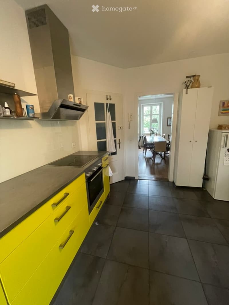 Modern kitchen with bright yellow cabinets, dark tiled floor, and stainless steel appliances. An open doorway leads to a dining area with a table and chairs.