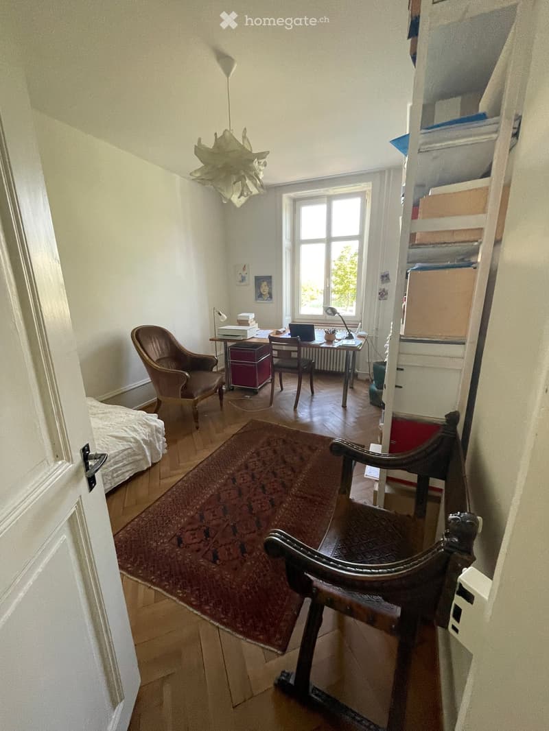 A furnished room with a wooden floor, a large window, a bed, a chair, a table with a laptop, a lamp, and a shelf with various items.