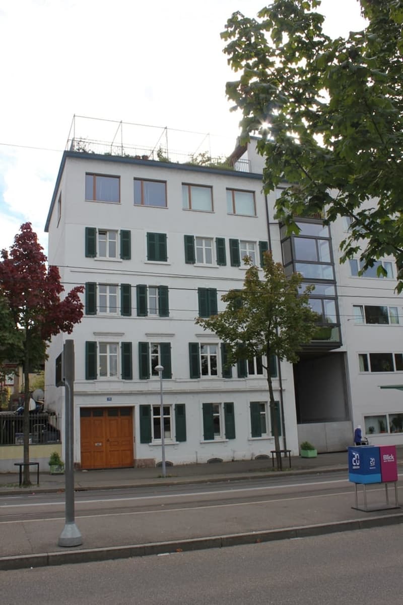A multi-story white building with green shutters and a wooden garage door, surrounded by trees and a sidewalk.