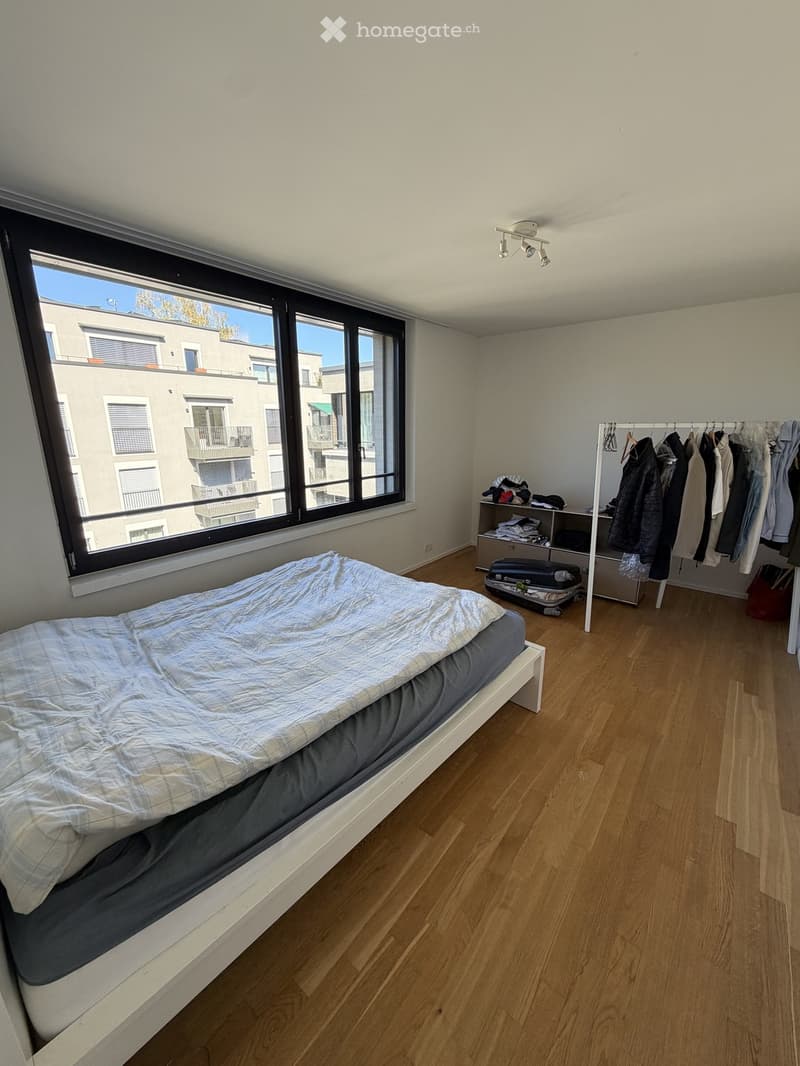 Bright bedroom with wooden flooring, a single bed with a grey duvet, a white wardrobe with hanging clothes, and a suitcase on the floor.