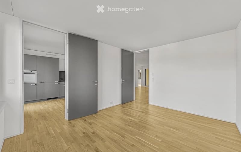 Empty room with light wooden flooring and white walls, featuring a closed door and an open doorway leading to another room with grey cabinets.