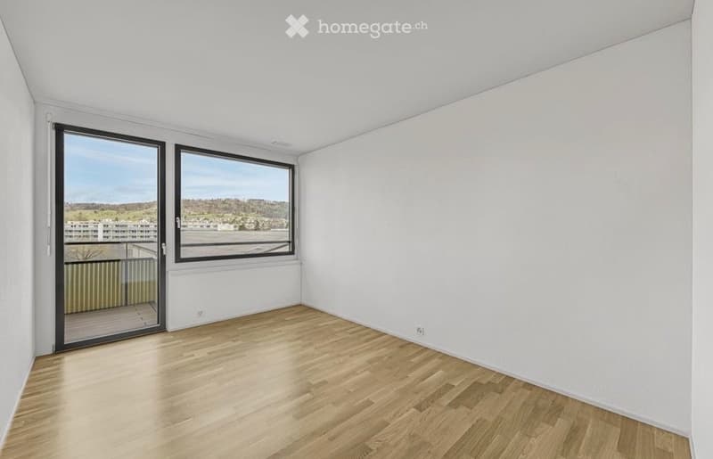 Empty room with wooden flooring, white walls, large windows, and a sliding glass door leading to a balcony with a scenic view.