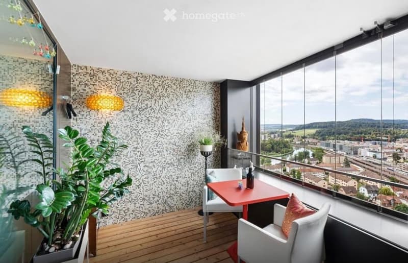 Modern balcony with wooden flooring, large windows offering a city view, a small red table with two white chairs, a potted plant, and decorative lighting.