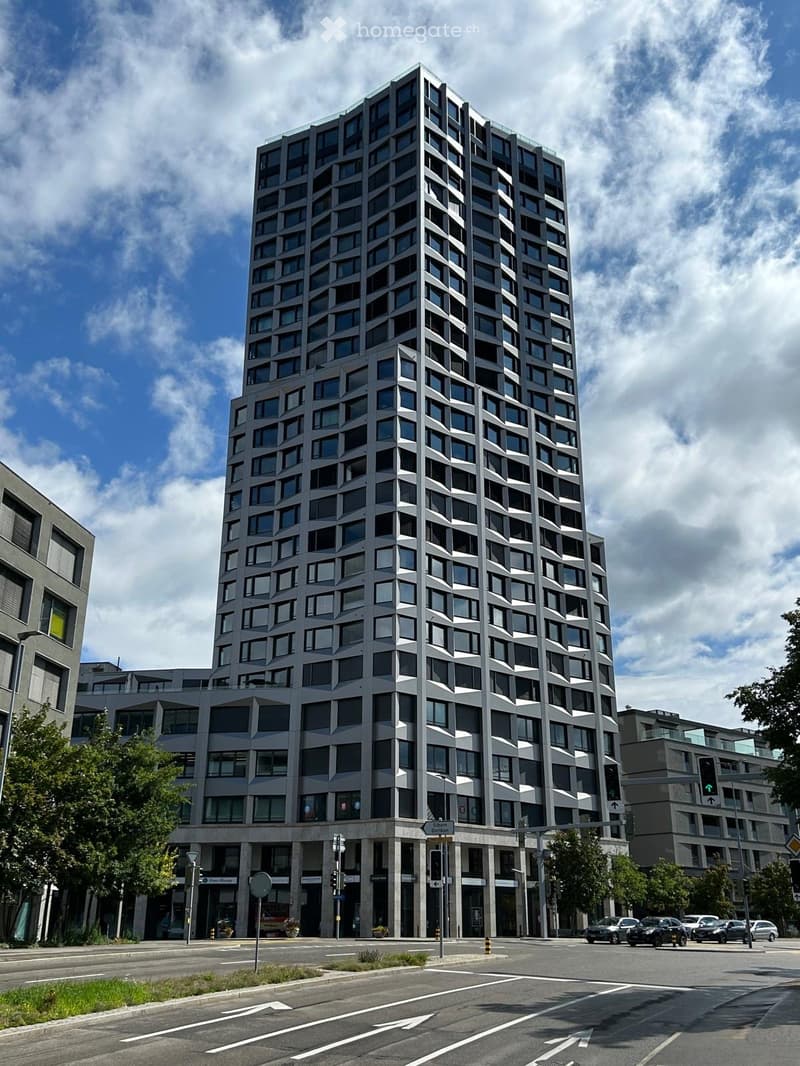 Modern high-rise building with a geometric facade, multiple floors, and numerous windows. The building is situated at a street corner with traffic signals and road markings visible.
