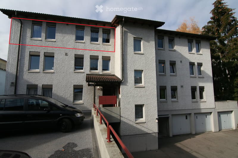 A two-story residential building with a textured light-colored facade, multiple windows, and a small entrance area with a covered walkway. A black van is parked in front of the building.