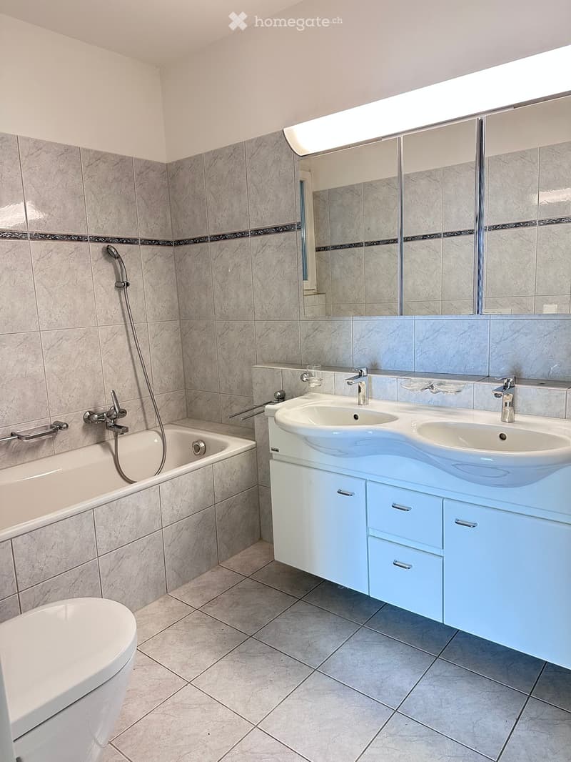 Modern bathroom with a double sink vanity, bathtub, toilet, and large mirror. Light blue cabinets and grey tiles.