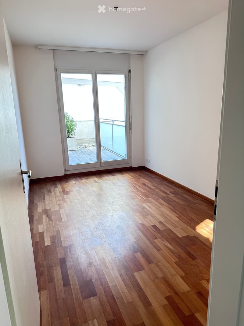 Empty room with wooden flooring, white walls, and a sliding glass door leading to a balcony with a plant visible outside.
