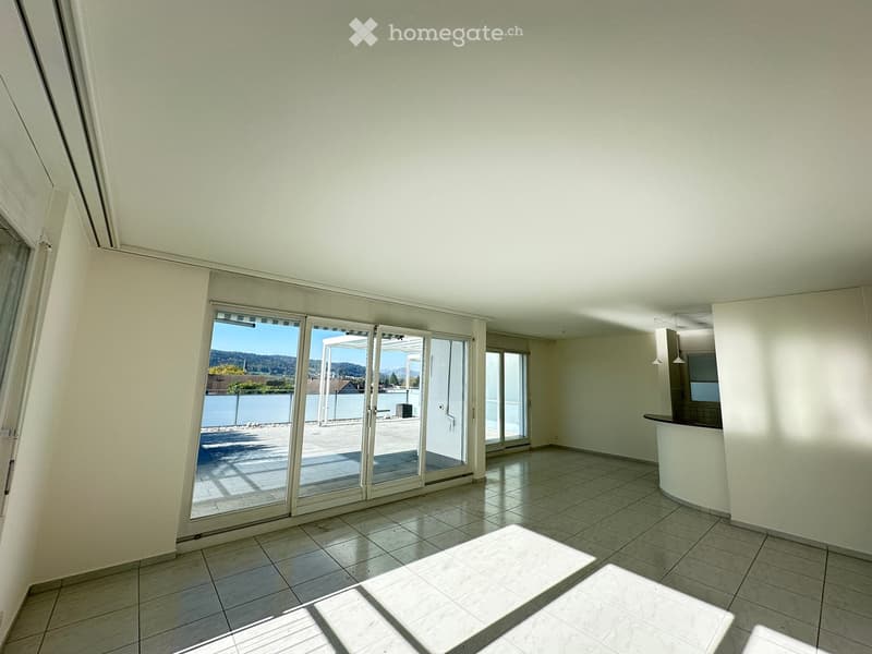 Empty room with tiled floor, large sliding glass doors leading to a balcony, and a small kitchen area visible in the background.