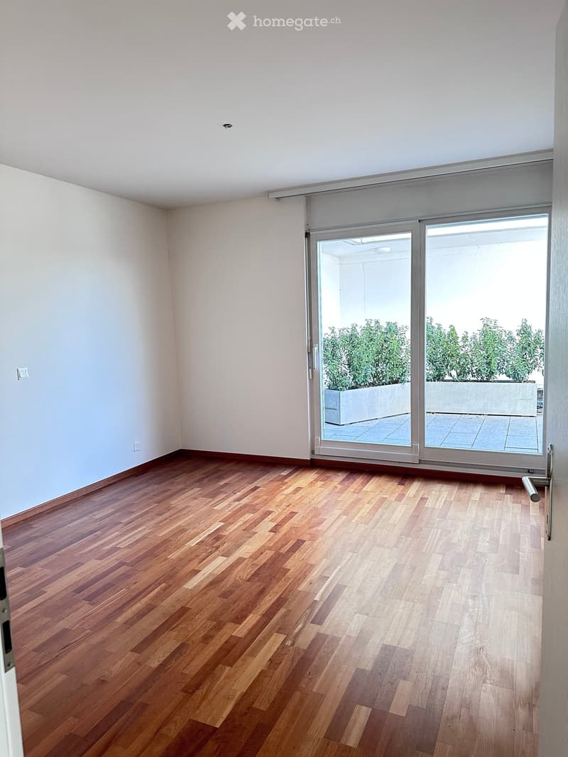 Empty room with wooden flooring and white walls, featuring a large sliding glass door leading to a balcony with potted plants.