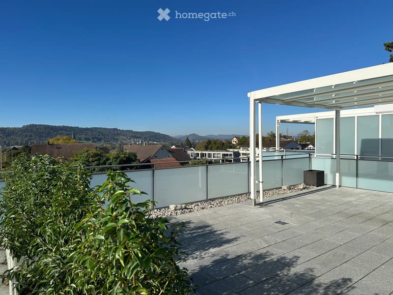 Modern balcony with glass railing, covered seating area, and scenic view of town and hills.