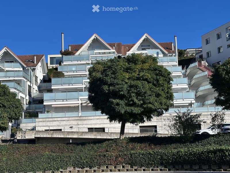 Modern multi-story residential building with balconies, surrounded by greenery and trees.