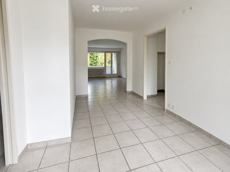 Empty hallway with white walls, tiled floor, and an arched opening leading to another room with large windows and a view of greenery.