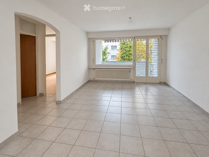 Empty room with tiled floor, white walls, large window with blinds, radiator, and an open doorway leading to another room.