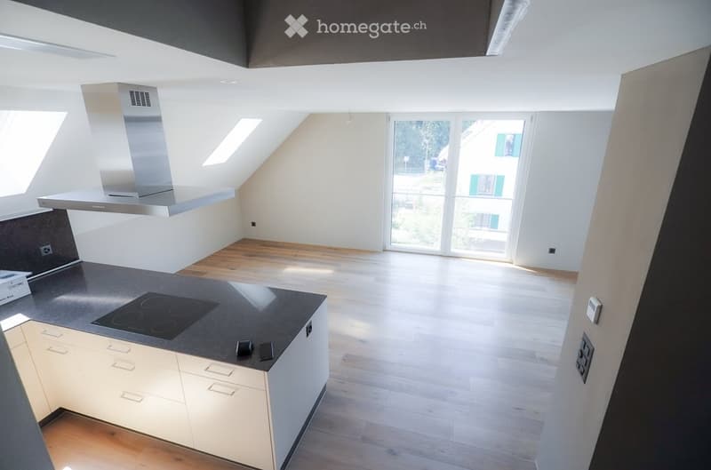 Open-plan attic space with wooden flooring, white walls, and a modern kitchen area featuring a built-in stove, countertop, and cabinets. Large windows and a sliding glass door provide natural light and access to the outdoor area.