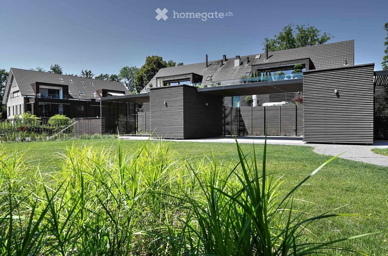 Modern two-story house with grey exterior, large windows, and a balcony on the upper level. The house is surrounded by a well-maintained lawn and trees.