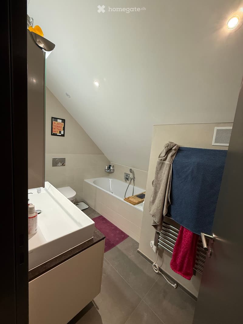 Modern bathroom with a sink, bathtub, toilet, towel rack, and towels. Features a sleek design with a wooden tray on the edge of the bathtub.